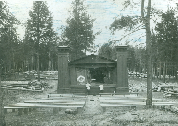 Ein altes Schwarz-Weiß-Foto einer Bühne im Wald mit zwei Personen davor, umgeben von Holzstämmen und Bäumen, mit dem Himmel im Hintergrund.