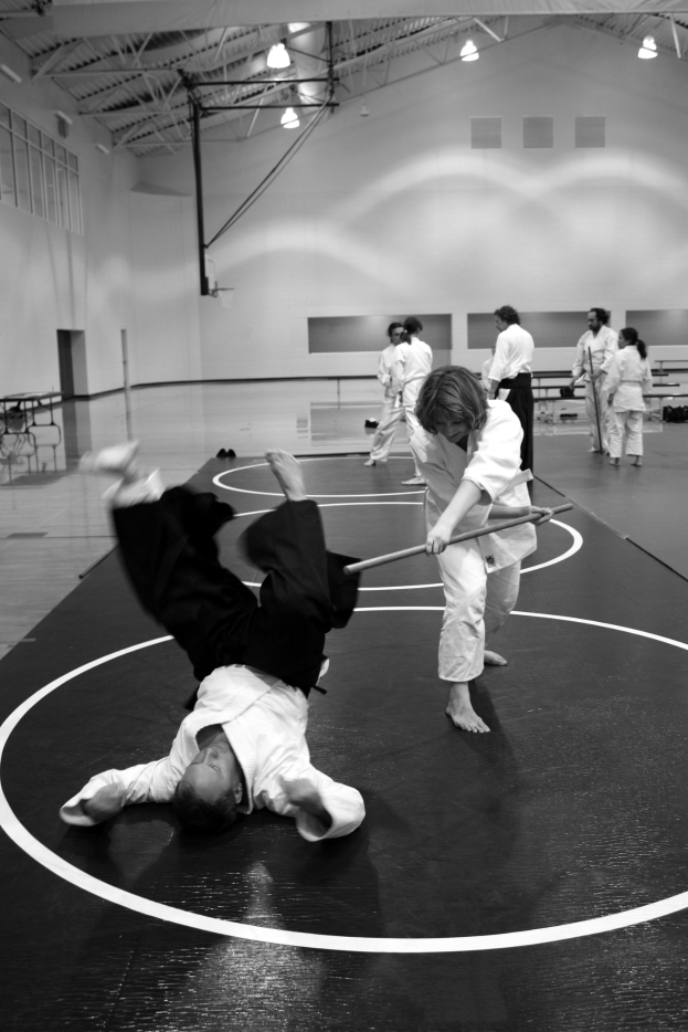 Zwei Personen in einer Karate-Halle üben Stockkampf, während andere zuschauen, mit Deckenbeleuchtung im Hintergrund.