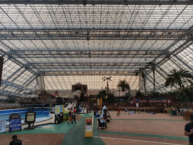 Gruppe von Menschen, die um ein Hallenbad in einem Sommerland-Freizeitpark herumstehen und sitzen, mit Bäumen, Informationsschildern und einem klaren blauen Himmel im Hintergrund.