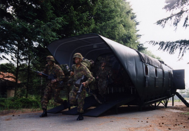Eine Gruppe von Soldaten in Helmen und mit Gewehren marschiert auf einer Schotterstraße neben einem Militärfahrzeug, mit Bäumen, Pflanzen, einem Zaun, einem Haus und einem klaren blauen Himmel im Hintergrund.