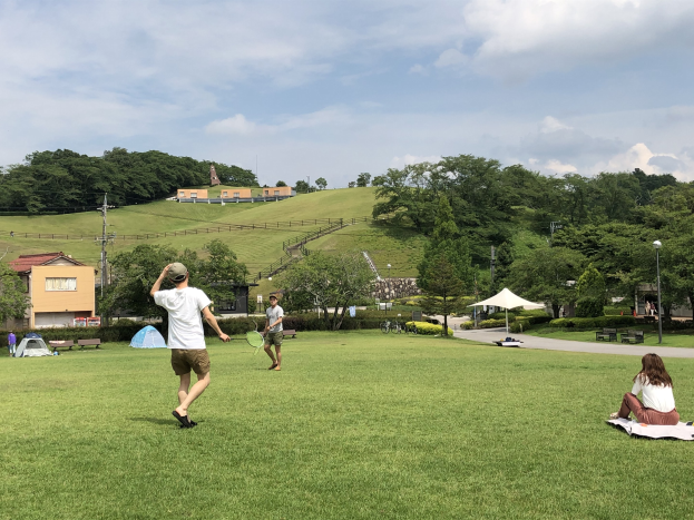 Gruppe von Menschen, die Badminton in einem Park spielt, mit Zelten, Straßenlaternen und Gebäuden im Hintergrund.