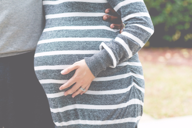 Schwangere Paare stehen zusammen mit Händen auf dem Bauch, tragen Schwangerschaftskleidung, umgeben von Bäumen.