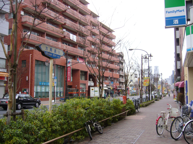 Eine Stadtstraße mit parkenden Fahrrädern, Gebäuden, Straßenlaternen, Verkehrsschildern, fahrenden Kraftfahrzeugen, Fußgängern auf dem Gehweg, Grünflächen und einem klaren blauen Himmel.