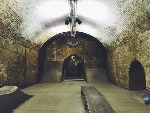 Ein Skatepark mit einem Backstein-Tunnel, einer Rampe, einer Bank und einem Schild, beleuchtet von Deckenlampen.