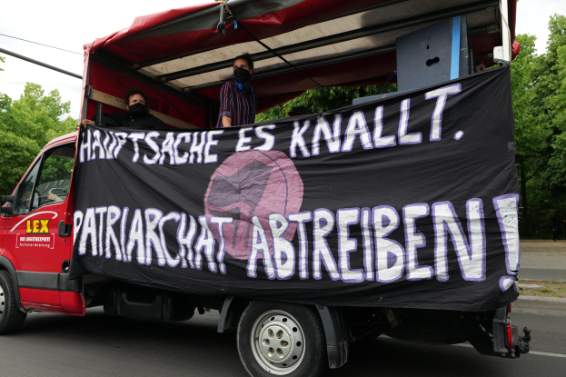 Roter Lkw mit einer Fahne, die eine Straße mit Bäumen an den Seiten entlangfährt, unter einem klaren blauen Himmel, zwei maskierte Personen sind im Inneren sichtbar.