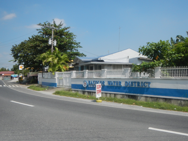 Straßenschild mit der Aufschrift "Wasserbezirk" neben einem Zaun, einer Wand, Bäumen, Häusern, Strommasten und einem bewölkten Himmel.