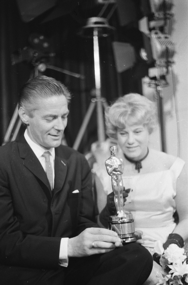 Ein Schwarz-Weiß-Bild eines formell gekleideten Mannes, der eine Oscar-Statue hält und eine Frau, die einen Blumenstrauß hält, nebeneinander sitzend mit Hintergrundbeleuchtung.