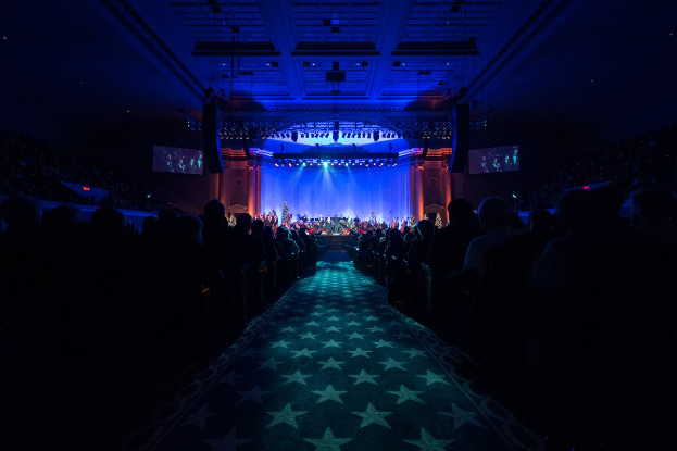 Ein großer Auditorium mit sitzenden Zuschauern, eine Bühne mit Musikern und über Kopfbeleuchtung und Bildschirme, was auf einen Konzertsaal während der Nationalkonvention 2019 hindeutet.
