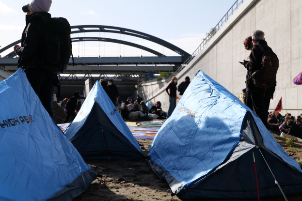 Gruppe von Menschen, die auf einem sandigen Strand in der Nähe von Zelten sitzen, mit einer Wand und einer Brücke im Hintergrund, die an einer Klimawandel-Demonstration teilnehmen, unter einem bewölkten Himmel.