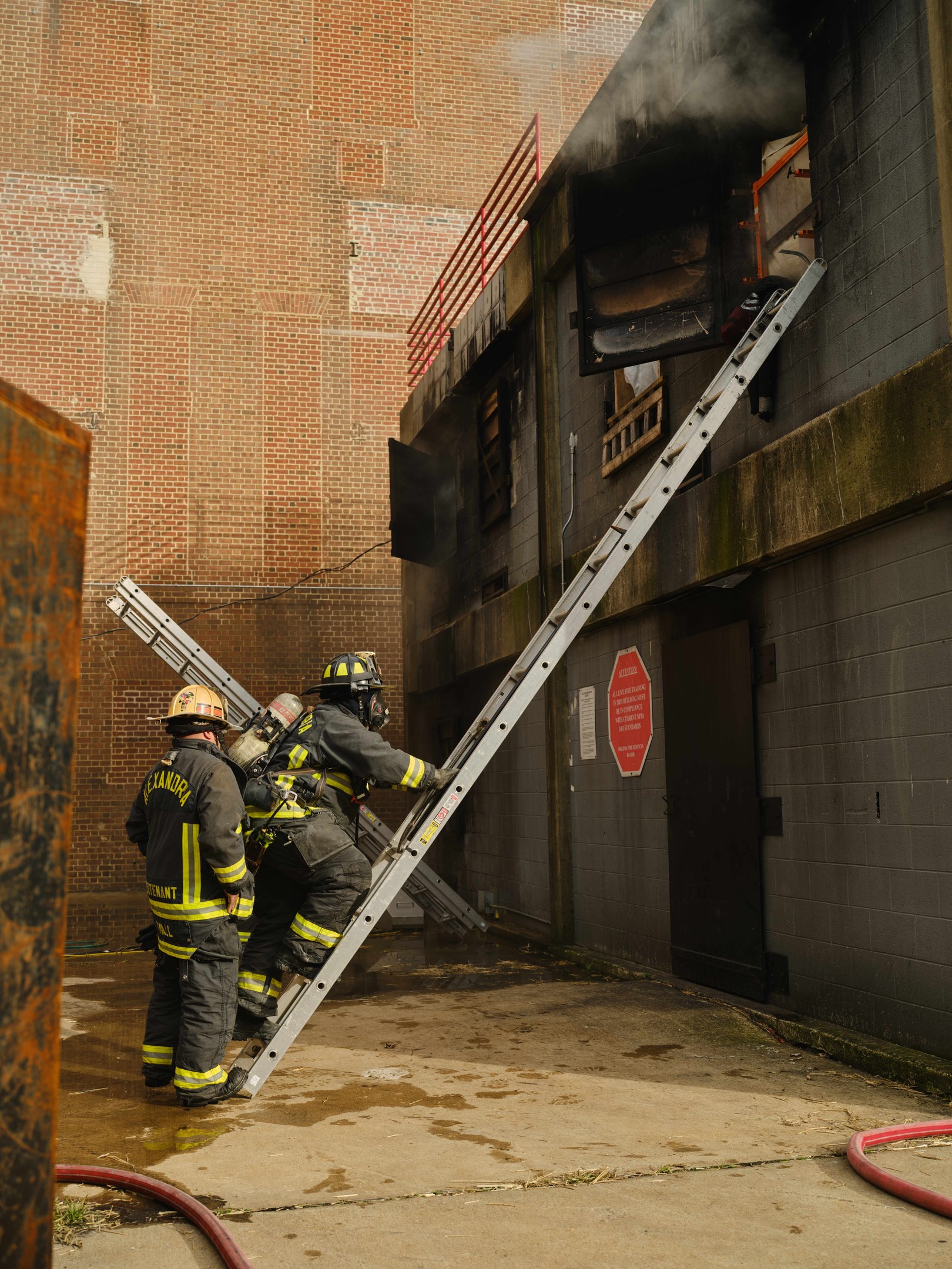 Feuerwehrleute in Helmen klettern an einer Leiter zu einem raucherfüllten Gebäude hinauf, mit einem metallischen Objekt links und Rohren an der Basis.