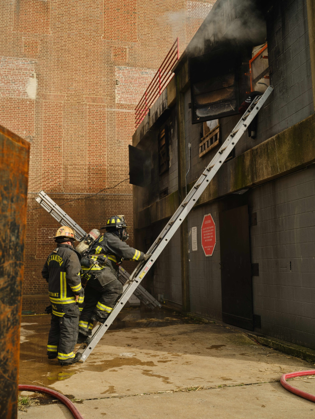 Feuerwehrleute in Helmen klettern an einer Leiter zu einem raucherfüllten Gebäude hinauf, mit einem metallischen Objekt links und Rohren an der Basis.