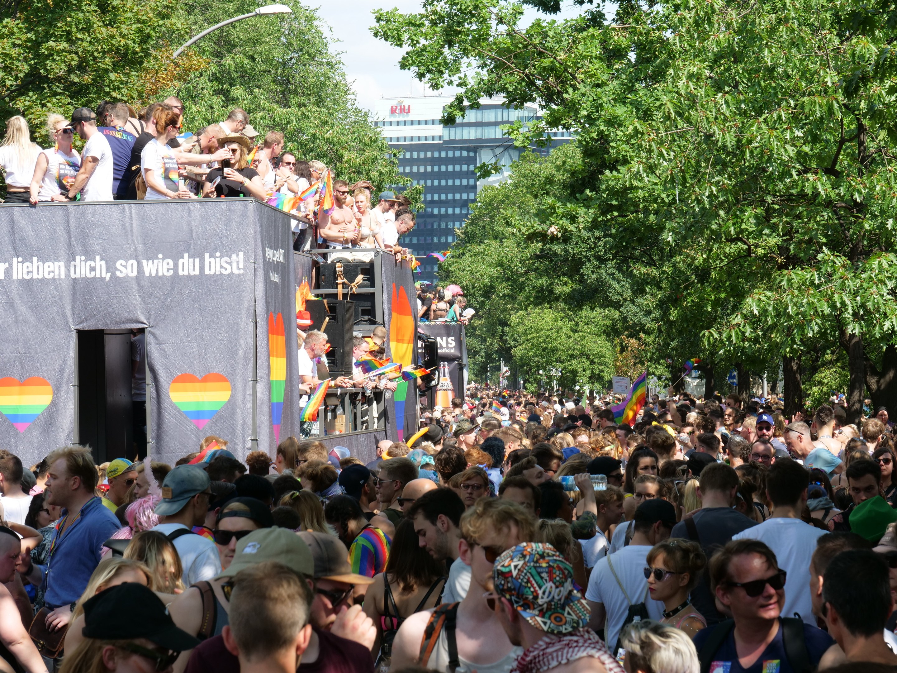 Große Menschenmenge bei der Berlin Pride Parade vor einem Lastwagen, viele tragen Mützen und Schutzbrillen, einige halten Fahnen, mit einem Banner auf dem Lastwagen und Bäumen, Gebäuden und einem Laternenpfahl im Hintergrund unter einem bewölkten Himmel.