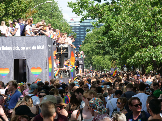 Große Menschenmenge bei der Berlin Pride Parade vor einem Lastwagen, viele tragen Mützen und Schutzbrillen, einige halten Fahnen, mit einem Banner auf dem Lastwagen und Bäumen, Gebäuden und einem Laternenpfahl im Hintergrund unter einem bewölkten Himmel.