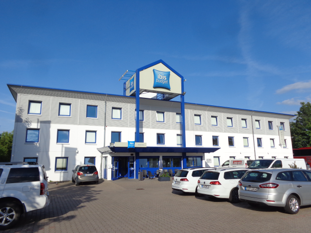 Ein Gebäude mit Fenstern und einem Schild mit der Aufschrift "ibis budget frankfurt airport", daneben geparkte Autos, Topfpflanzen, ein Straßenmast, Bäume und ein bewölkter Himmel.