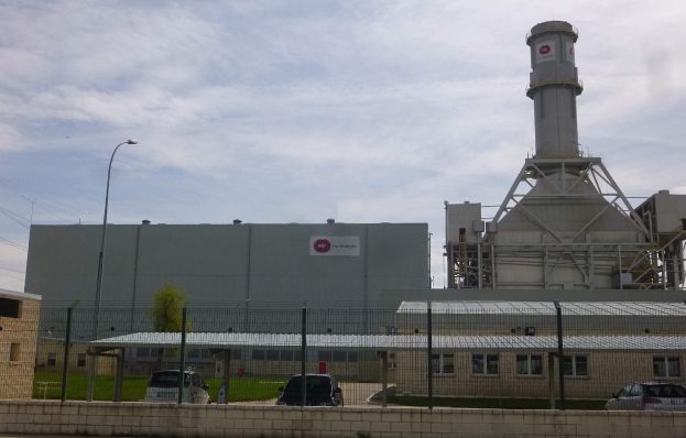 Großes Industriegebäude mit einem zentralen Turm, umgeben von einem Zaun, Fahrzeugen auf der Straße davor, Strommasten, Straßenlaternen, Drähte, Bäume, Gras und ein bewölkter Himmel.