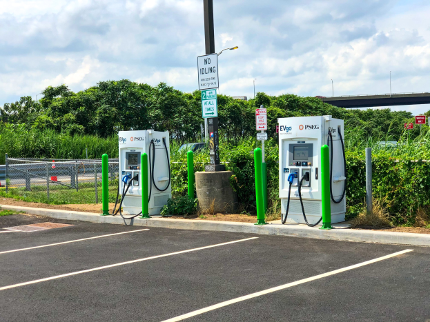 Elektrofahrzeug-Ladestation auf einem Parkplatz mit umgebender Infrastruktur, Vegetation und einer Brücke im Hintergrund bei bewölktem Himmel.