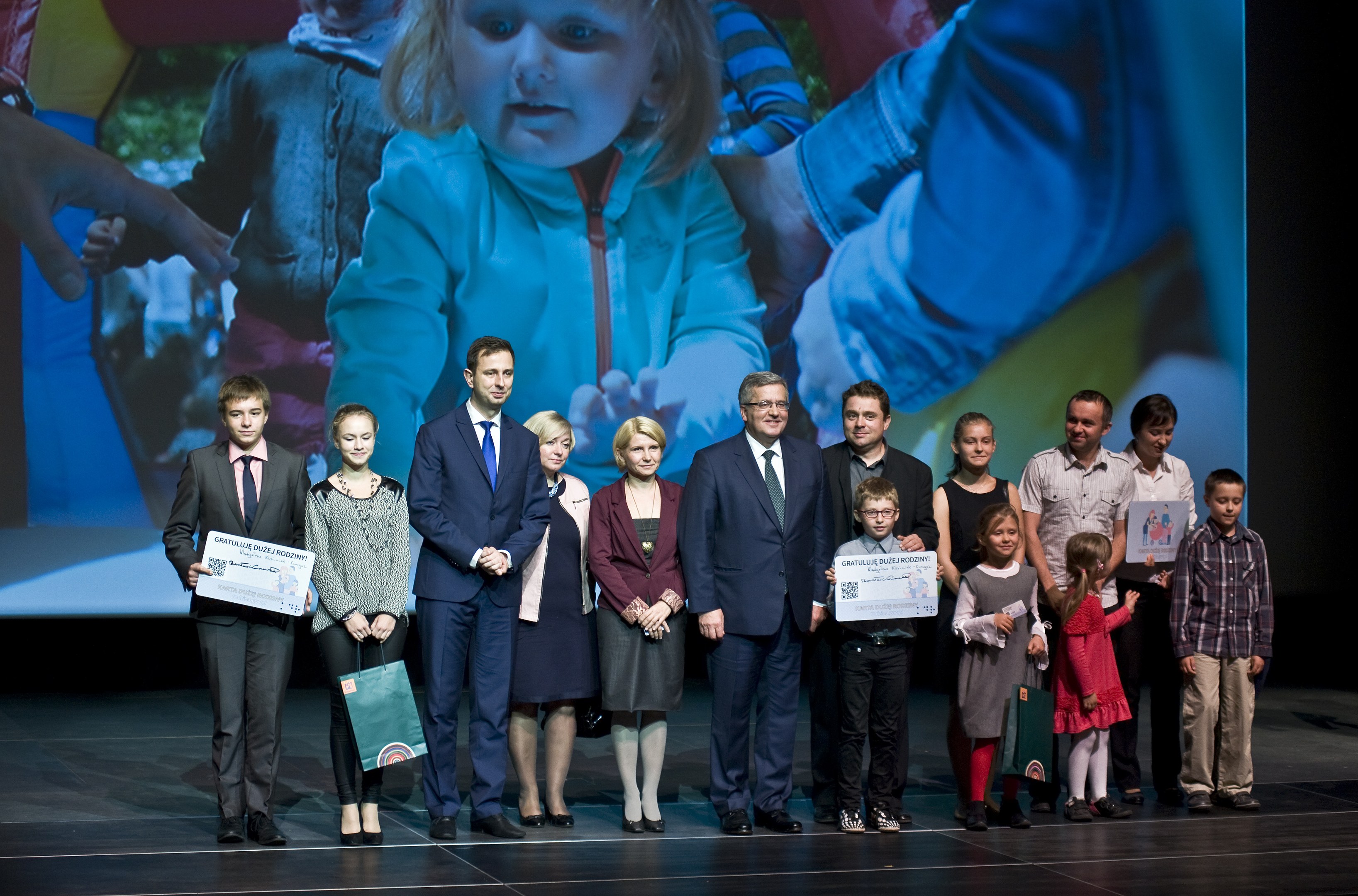 Gruppe von Menschen auf einer Buhne mit Taschen und Urkunden, im Hintergrund ein Bildschirm mit einem Kind und anderen.