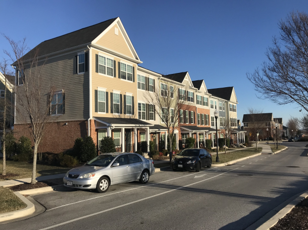 Eine Reihe von Stadthäusern mit parkenden Autos, Bäumen und Laternen in einer Wohngegend unter einem sichtbaren Himmel.