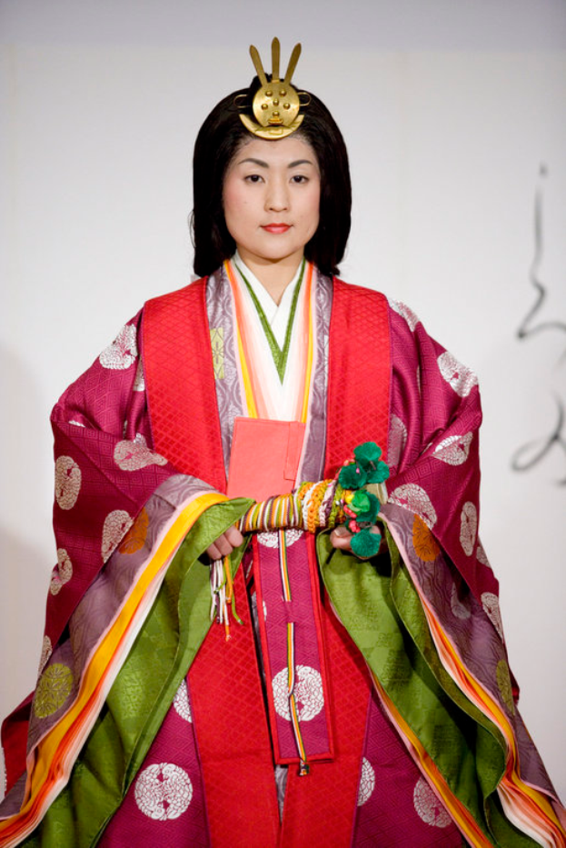 Eine Frau in einem traditionellen japanischen Kimono, mit einem leuchtend roten und grünen Kleid mit aufwändigen Mustern, steht selbstbewusst auf einem Laufsteg, ihr Haar ordentlich zu einem Knoten gebunden.