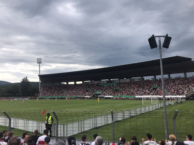 Eine große Menschenmenge schaut einem Fußballspiel in einem eingezäunten Stadion unter beleuchteten Masten zu, mit Bäumen und Hügeln im Hintergrund unter einem bewölkten Himmel.