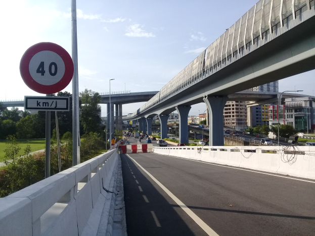 Autobahn mit Geschwindigkeitsbegrenzungsschild, Fahrzeugen, Brückenpfeilern, Laternen, B├Ąumen, Geb├Ąuden und bew├Âlktem Himmel im Hintergrund.
