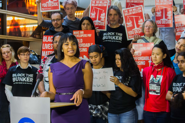 Eine Black Lives Matter-Aktivistin, die vor einer Menge mit Schildern an einem Rednerpult mit Mikrofon steht, vor Lichtern und anderen Gegenständen.