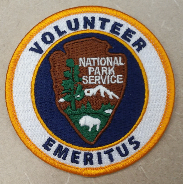 Ein rundes Patch mit blauem Rand und weißem Hintergrund, das schwarze Text 'Volunteer National Park Service Emeritus' in der Mitte eines gelben Kreises zeigt.