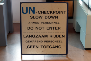 Ein Schild auf dem Boden lautet "UN-Checkpoint Slow Down Armed Personnel - Do Not Enter", mit einem Glastisch mit verschiedenen Gegenständen darauf zur Rechten und einer Wand im Hintergrund.