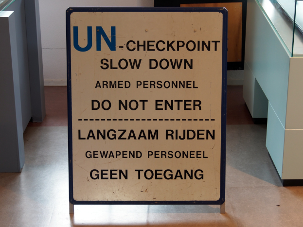 Ein Schild auf dem Boden lautet "UN-Checkpoint Slow Down Armed Personnel - Do Not Enter", mit einem Glastisch mit verschiedenen Gegenständen darauf zur Rechten und einer Wand im Hintergrund.