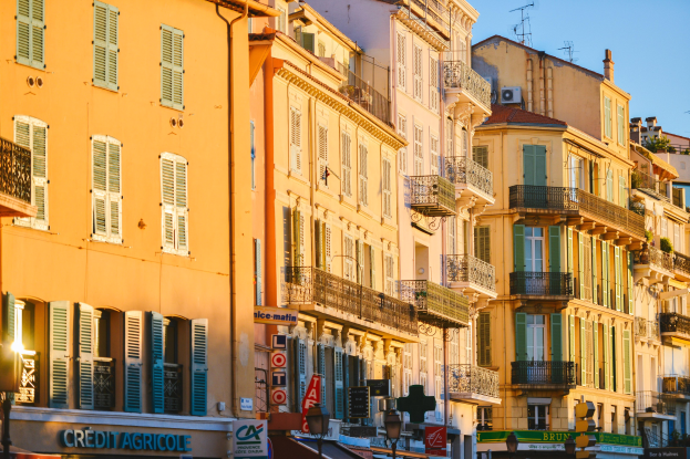 Eine Stadtstraße mit hohen Gebäuden, die Fenster, Balkone und Schilder aufweist und von Bäumen und Straßenlaternen gesäumt ist, unter einem klaren blauen Himmel.