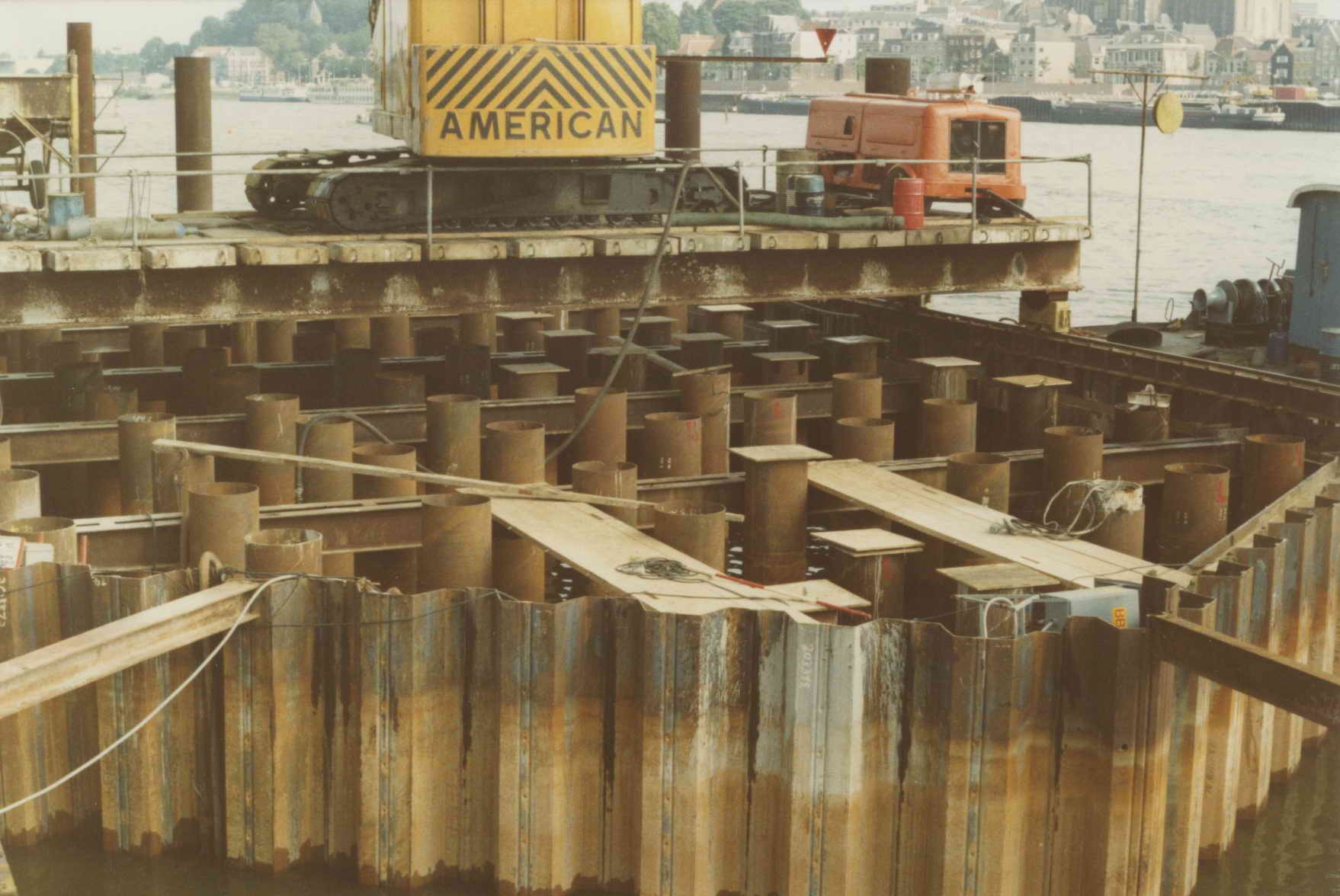 Baustelle mit einem Kran, der Stahlstangen von einem Haufen hebt, umgeben von Wasser, Gebäuden, Bäumen und einem klaren blauen Himmel.