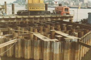 Baustelle mit einem Kran, der Stahlstangen von einem Haufen hebt, umgeben von Wasser, Gebäuden, Bäumen und einem klaren blauen Himmel.