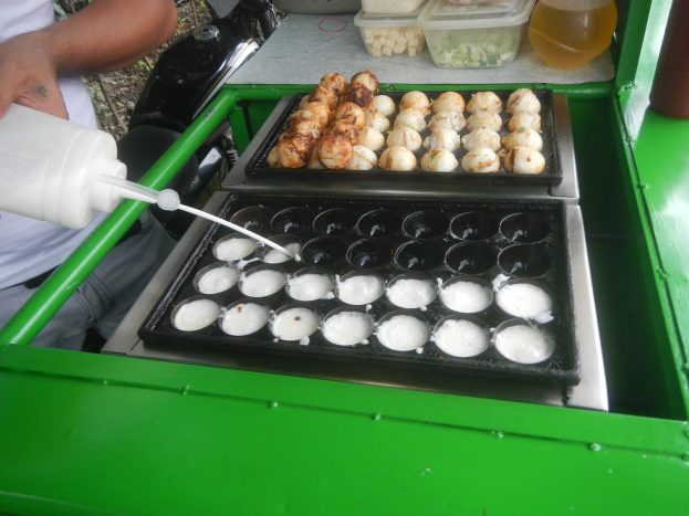 Ein Mann gießt Milch aus einer Flasche in eine Schüssel mit Essen auf einem grünen Tisch, umgeben von Schüsseln, Flaschen und anderen Gegenständen.