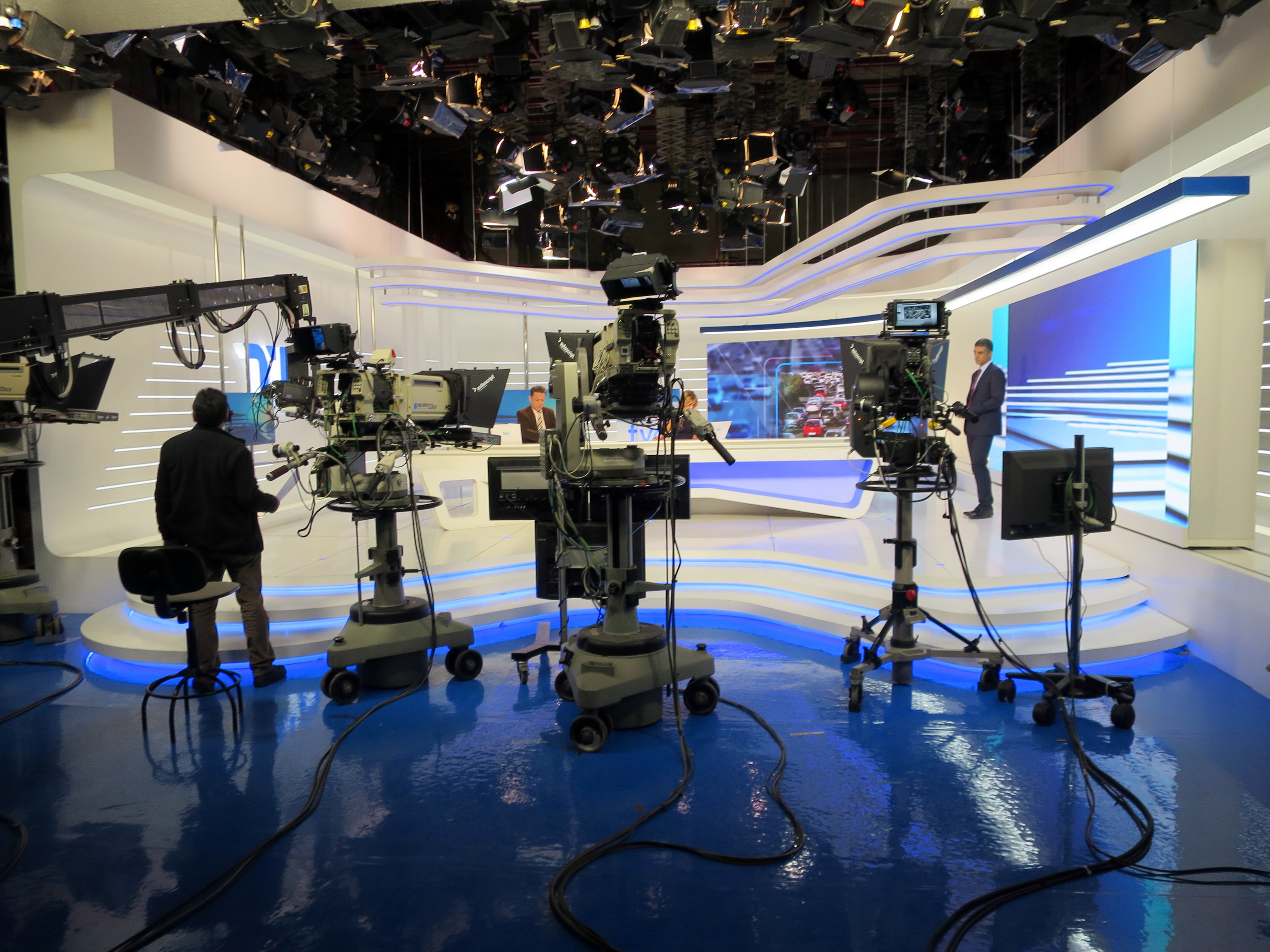 Ein TV-Studio mit mehreren Kameras auf dem Boden, einer sitzenden Person im Vordergrund und einigen stehenden Personen im Hintergrund, mit an der Decke montierten Scheinwerfern für eine Livesendung.