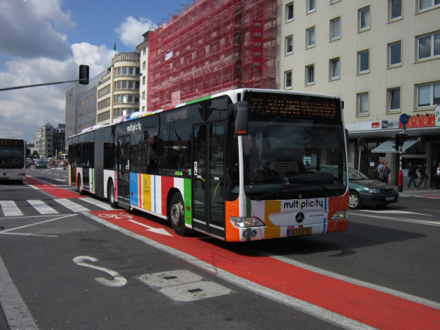 Eine Stadtbus fährt auf einer Straße mit hohen Gebäuden entlang, mit Menschen auf dem Gehweg, Ampeln und Texttafeln, unter einem bewölktem Himmel.
