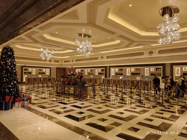 Ein geräumiger Eingangsbereich mit einem zentralen Weihnachtsbaum, umgeben von Geschenkboxen und festlichen Dekorationen, mit Menschen, Absperrpollern mit Seilen, Tischen mit Blumenbouquets und gerahmten Fotos an den Wänden, unter einer Decke mit Lichtern und Kronleuchtern.