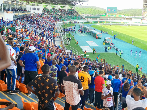 Eine große Menschenmenge steht und sitzt vor einem eingezäunten Fußballfeld mit Hügeln und einem klaren blauen Himmel im Hintergrund während der 2019 African Cup of Nations.