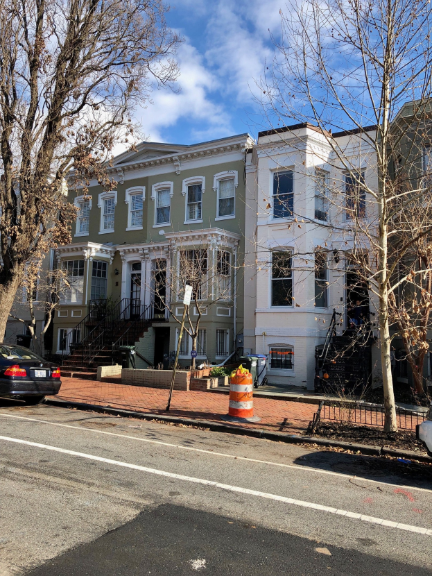 Eine Reihe von Stadthäusern an einer Straßenecke in Washington, DC, mit Fahrzeugen, einer Verkehrskegel, Bäumen und Gebäuden mit Fenstern und Treppen mit Geländern unter einem bewölkten Himmel.