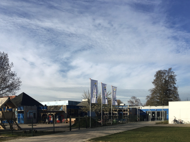 Großes Gebäude mit Metallzaun, Weg, Bäumen, Bannern, einem geparkten Fahrrad, nahen Häusern und einem bewölkten Himmel, identifiziert als der neue Spielbereich für Kinder an der Schule.