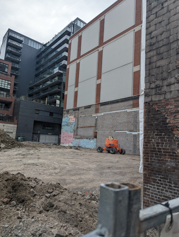 Baustelle mit einem teilweise abgerissenen Gebäude im Hintergrund, ein Fahrzeug am Boden, ein Metallzaun und ein bewölkter Himmel.