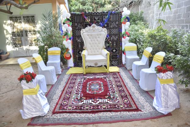 Eine Gartenhochzeit mit weißen Stühlen, einem roten Teppich, Blumenbouquets, Dekorationselementen, Luftballons und umgebender Vegetation in der Nähe eines Gebäudes mit Fenstern und einer Tür.