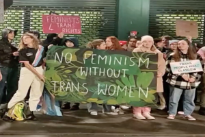 Eine Gruppe von Menschen hält ein Transparent mit der Aufschrift "Kein Feminismus ohne Trans-Frauen" und verschiedene Schilder in der Öffentlichkeit.