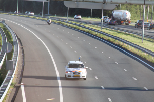 Ein Auto auf einer Autobahn mit einem stationären Polizeiwagen am Rande, andere Fahrzeuge und Bäume sowie Straßenstrukturen im Hintergrund.