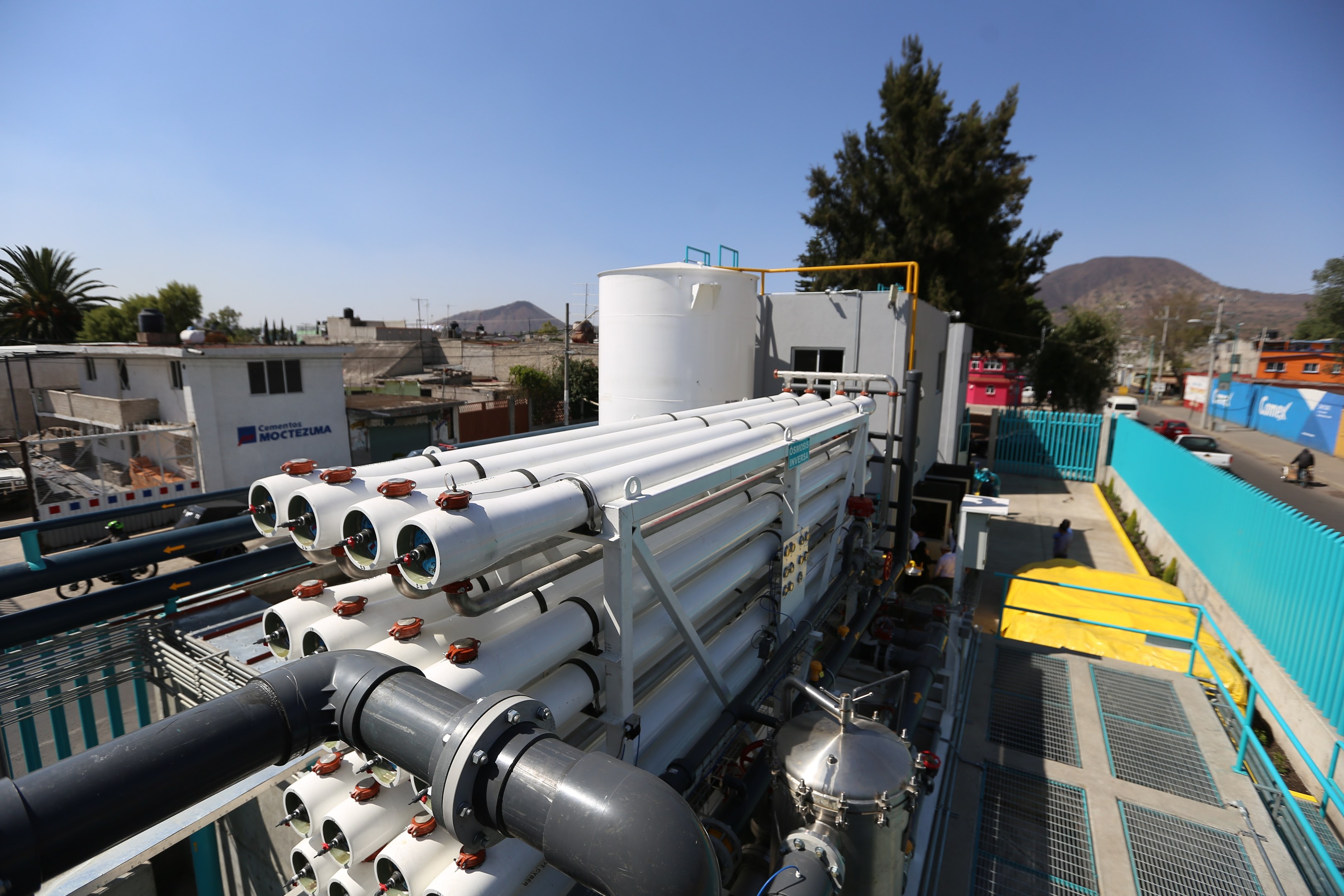 Große Gruppe von Rohren auf dem Dach eines Gebäudes mit umgebenen Industriegeräten, Fahrzeugen, Menschen und städtischer Landschaft unter einem klaren blauen Himmel.