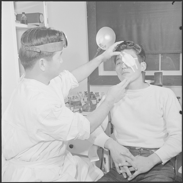 Schwarz-weiß-Foto eines Mannes bei einer Augenuntersuchung durch einen Arzt, mit medizinischen Geräten und einem Fenster mit Vorhang im Hintergrund.