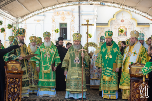 Eine Gruppe von Priestern steht vor einer Kirche, wobei einer ein Buch und ein Mikrofon auf der linken Seite hält, und ein Kreuz, Blumen und ein Gebäude im Hintergrund zu sehen sind.