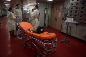 Eine Gruppe von Menschen in Schutzausrüstung steht um eine Trage in einem Krankenhauszimmer mit medizinischer Ausrüstung, darunter eine Trage mit Tasche und anderen Gegenständen, ein Ständer mit verschiedenen Gegenständen, eine Tür, eine Säule, ein Schild und eine Decke mit Lampen.