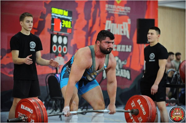 Ein Mann, der eine Hantel in einem Gewichtheben-Wettbewerb hochhebt, umgeben von Zuschauern, mit Wettkampfausrüstung und Hinweisschildern, die die 2018 World Weightlifting Championships anzeigen.