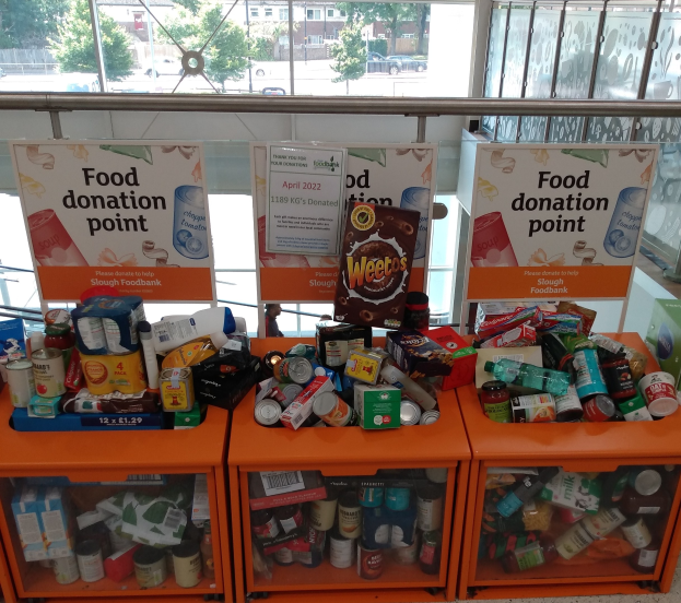 Orange Schränke mit Lebensmittelspenden, darunter Flaschen, Dosen und Kartons, mit Informationsschildern und Blick auf Fahrzeuge, Bäume, Gebäude und Himmel durch Glasfenster.