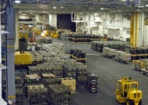 Großes Lagerinnere mit mehreren Lkws, Containern, Menschen, Deckenbeleuchtung, Metallstangen und einem sichtbaren Rad.